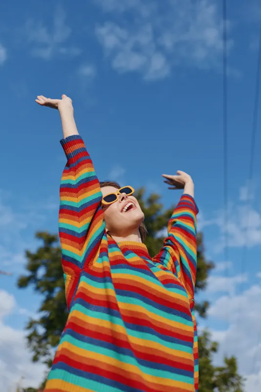 Woman in a rainbow sweater and sunglasses, joyful young adult smiling with arms raised outdoors under blue sky, casual outfit and carefree vibe for lifestyle, travel and fashion scenes.