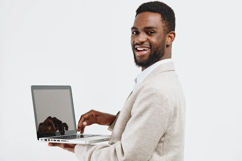 Happy smiling man with dark skin holding a laptop computer and pointing at the screen isolated on plain white background. Portrait of a cheerful person using technology, casual clothing, modern concep