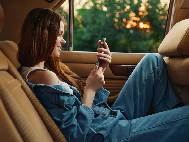Young woman in denim overalls enjoying her smartphone in a car during sunset, embodying relaxation and modern lifestyle.