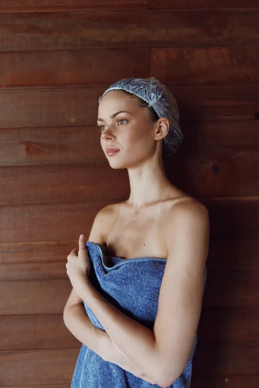 young woman wrapped in blue towel with hair net stands against wooden wall, natural light highlighting serene expression and calm atmosphere