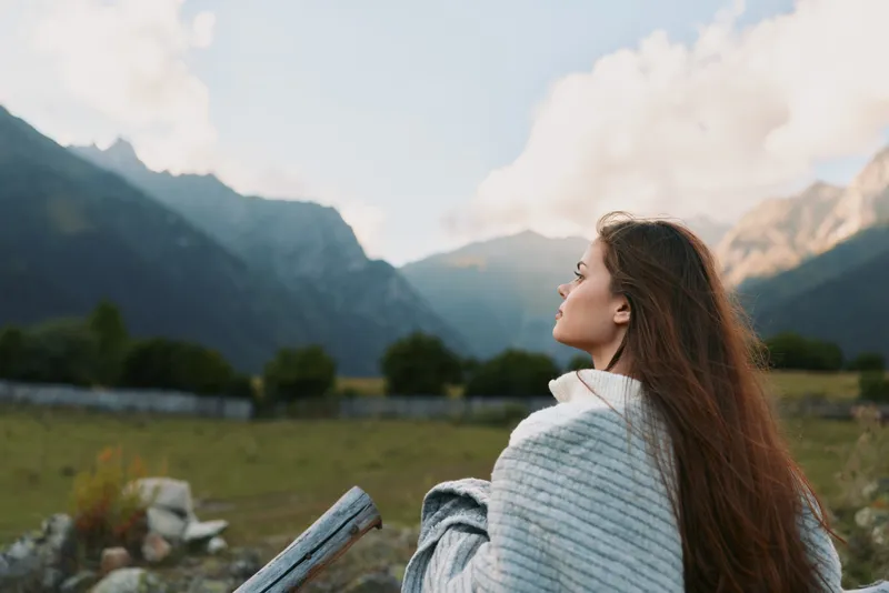 Young woman wrapped in blanket looking at mountain landscape thoughtful mood outdoor natural light peaceful atmosphere lifestyle concept