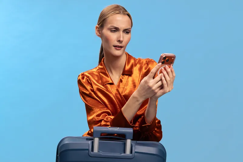 Young woman in orange satin blouse looks at smartphone while leaning on blue suitcase. Studio portrait on blue background, concept of travel, communication, and modern lifestyle.