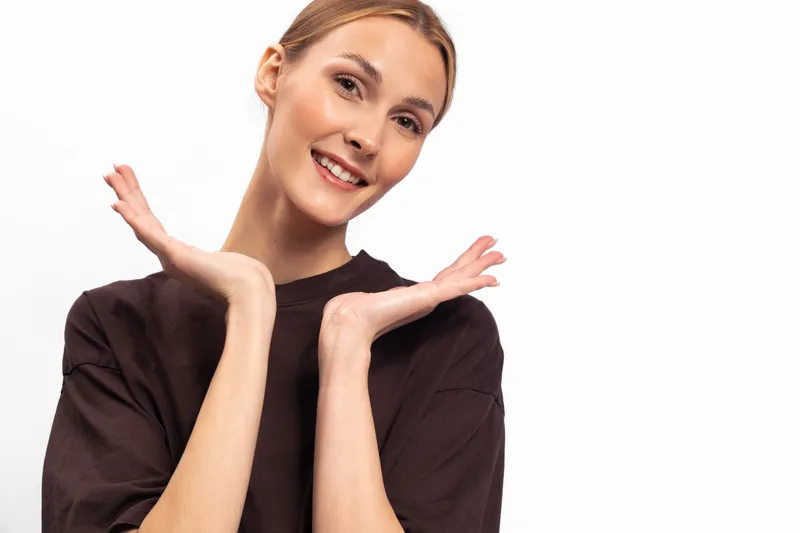 Smiling young woman with natural makeup and brown hair wearing dark shirt poses with hands near face on white background. Concept of happiness, youth, and casual style.