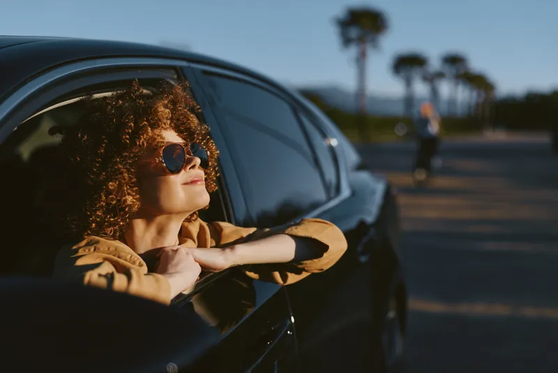 Smiling woman with curly hair leaning on a car window. Retro film color aesthetic on a sunny road. Lifestyle, travel, and carefree confidence captured with warm, soft lighting.