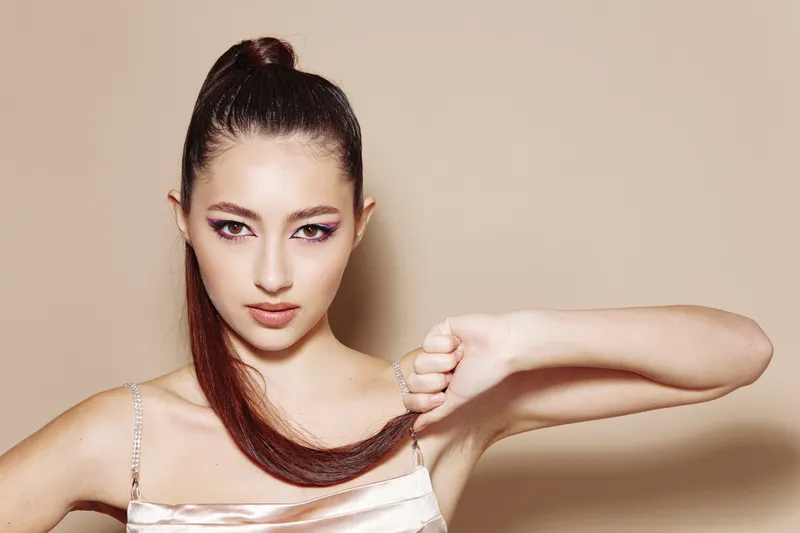 Young woman with sleek high ponytail firted dress poses holding hair. Her focused expression highlights unique beauty fashion and creative style in studio. Beige background complements soft tones.