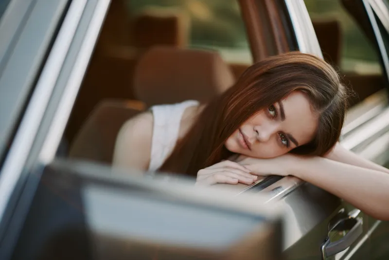 Young woman with long brown hair leaning out of a car window, expressing a feeling of longing in a serene outdoor setting during sunset.