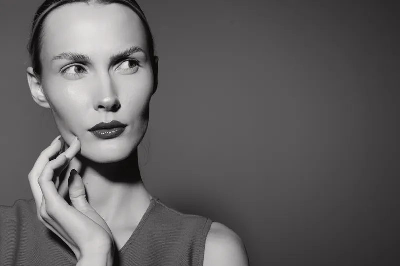 Portrait of a slender Caucasian woman with fair skin and red lips looking to the side, hand near face. Black and white studio photo with dramatic lighting, fashion concept.