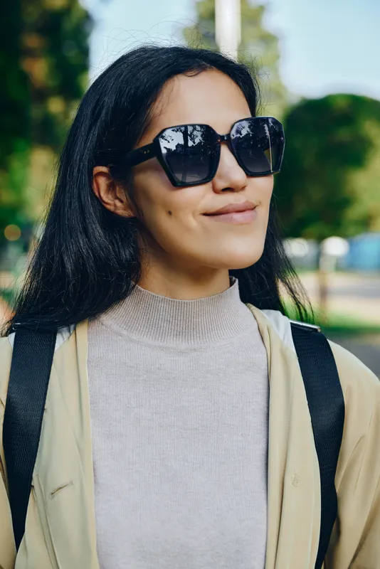 Sunglasses, woman outdoors in park wearing a chic outfit, sunny daylight, fashion vibe, subtle smile, casual lifestyle scene with backpack and stylish accessory
