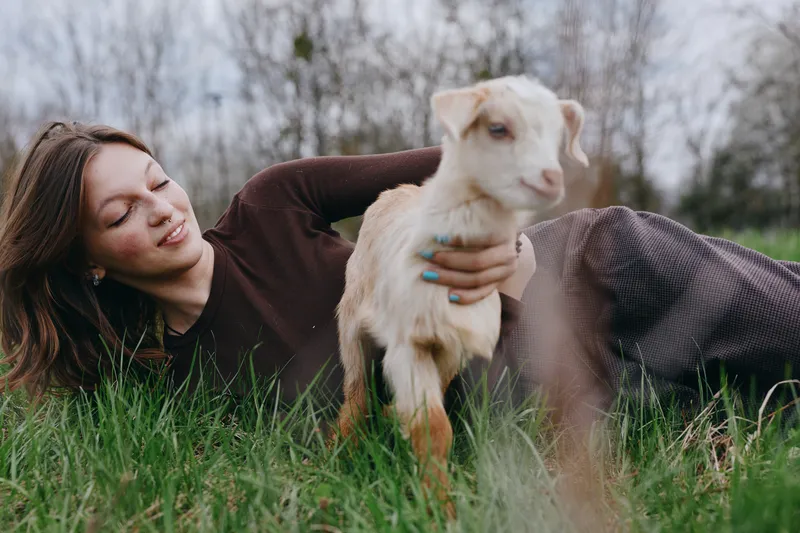 Cheerful woman outdoors, lying on grass with a goat kid nearby, warm sunlight highlights a relaxed lifestyle moment, friendly nature scene, candid interaction and calm bond between animal and human