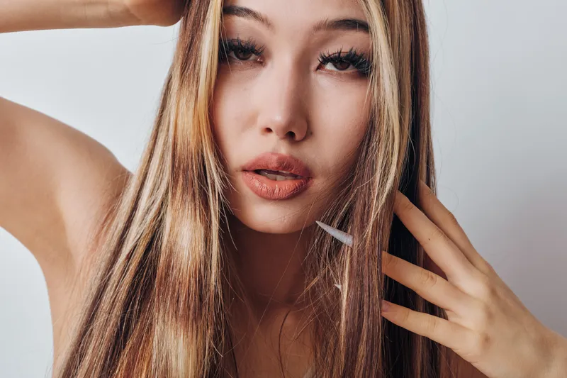 Woman with long blonde hair posing closeup for beauty portrait. Female model with natural makeup and hand near face against white background in studio lighting.