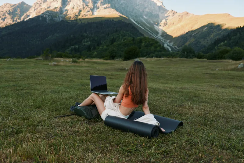 A woman sits on a grassy field with a laptop, enjoying outdoor work in a scenic mountainside setting. She looks relaxed and focused, creating a calm, productive atmosphere amid nature.