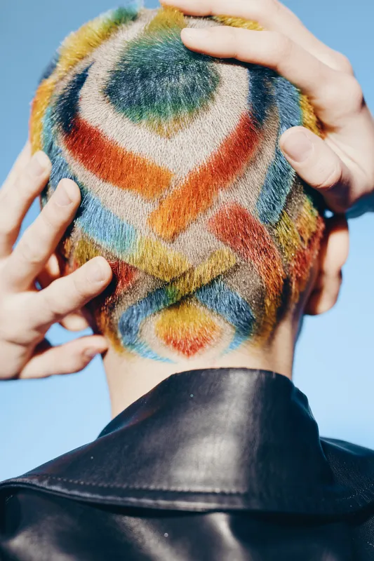 Creative woman with colorful dyed buzz cut viewed from behind, vibrant hair patterns and black leather jacket under clear blue sky, artistic urban style.