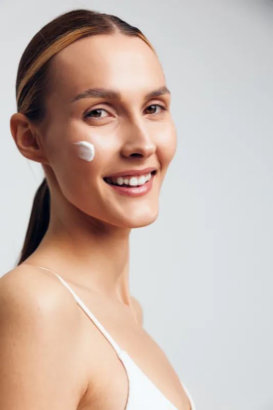 Smiling woman with clear skin applying cream on cheek, natural beauty concept, fresh face, minimal makeup, soft lighting, clean background, skincare routine, healthy lifestyle