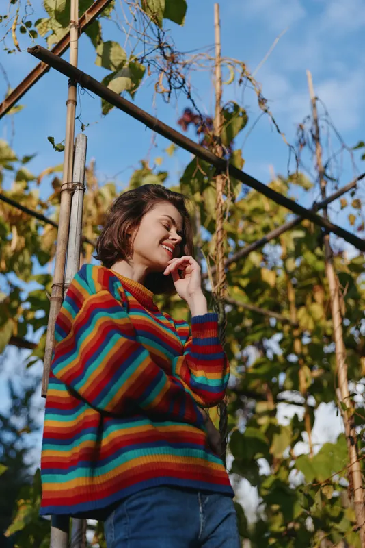 Woman in her mid twenties in a striped sweater smiling in a garden by a wooden trellis, casual outfit and brown hair, relaxed pose with warm natural light for lifestyle portrait outdoors.