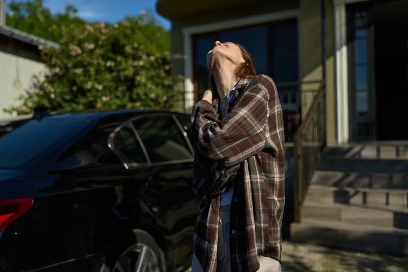 Young woman outdoors beside a car wears a plaid coat tilting back her head to enjoy sunlight and a cool breeze in a sunlit driveway with soft shadows and a relaxed mood