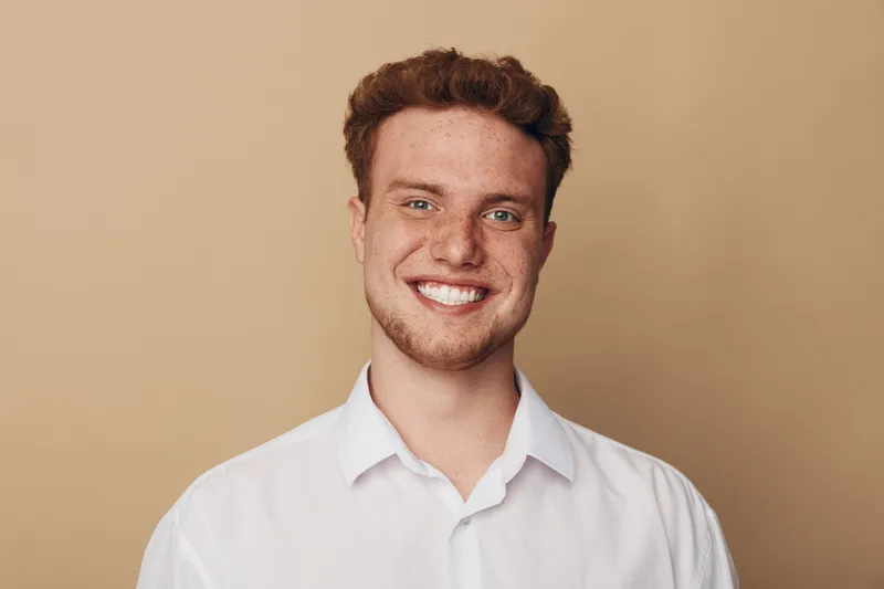 Smiling young man with curly hair wearing white shirt looks at camera on beige background. Concept of happiness, confidence, youth, and casual portrait photography.