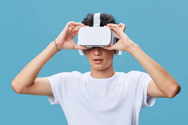 Young man wearing virtual reality headset and headphones against blue background, enjoying immersive technology experience in casual white t-shirt.