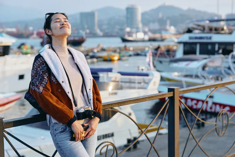 Woman leaning on railing at harbor, warm sunlight bathes boats and water, casual fashion with scarf and coat, serene travel mood, seaside breeze and distant skyline in view.