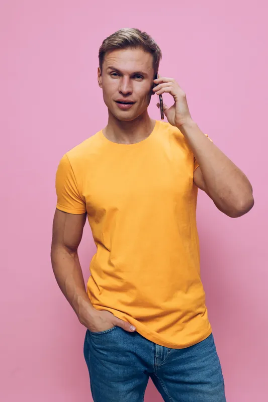 Full body confident man talking on mobile phone wearing casual yellow t-shirt and blue jeans smiles looks relaxed isolated on pink background studio portrait. People lifestyle concept