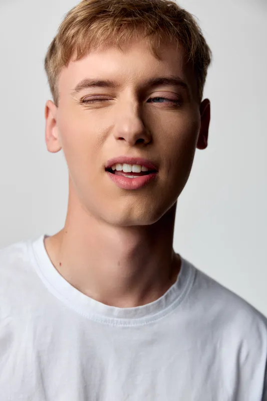 Winking man with casual expression wearing a white tshirt, closeup face, cheeky mood and playful gesture captured in subtle studio lighting for authentic lifestyle appeal