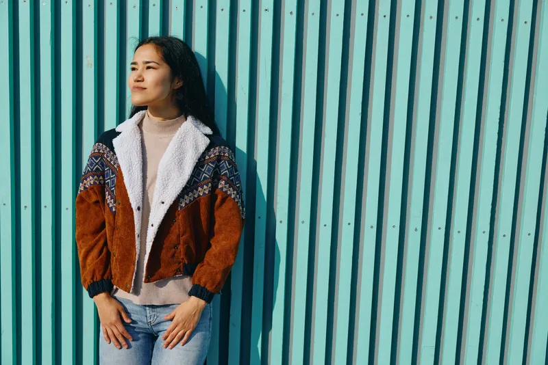Woman leaning against a teal striped wall, wearing a colorblocked jacket and knit sweater, casual street style in an urban setting with warm daylight