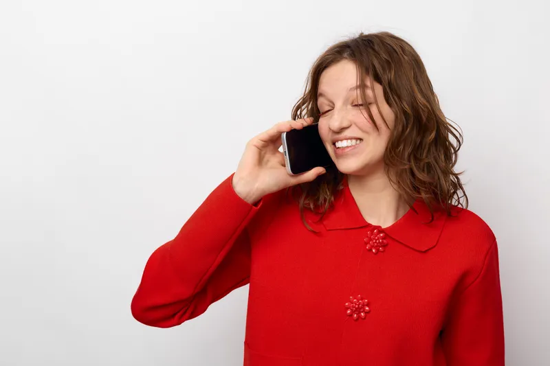 Young woman in red sweater talking on smartphone with eyes closed and smiling. Isolated on white background. Concept of communication, happiness, casual lifestyle, and technology use.