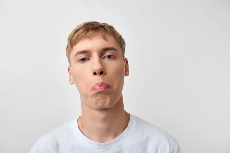 Young man with a pouty face and puzzled expression, close up shot, casual vibe, white tee, simple studio lighting, humorous mood and candid style vibe
