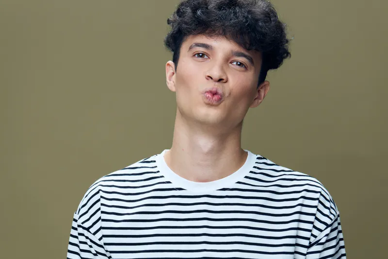 young man with curly hair wearing striped shirt making kissy face with green background expressing playful and fun mood