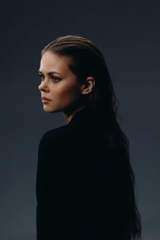 Serious woman with braided hair wearing black clothes against dark background in moody lighting.