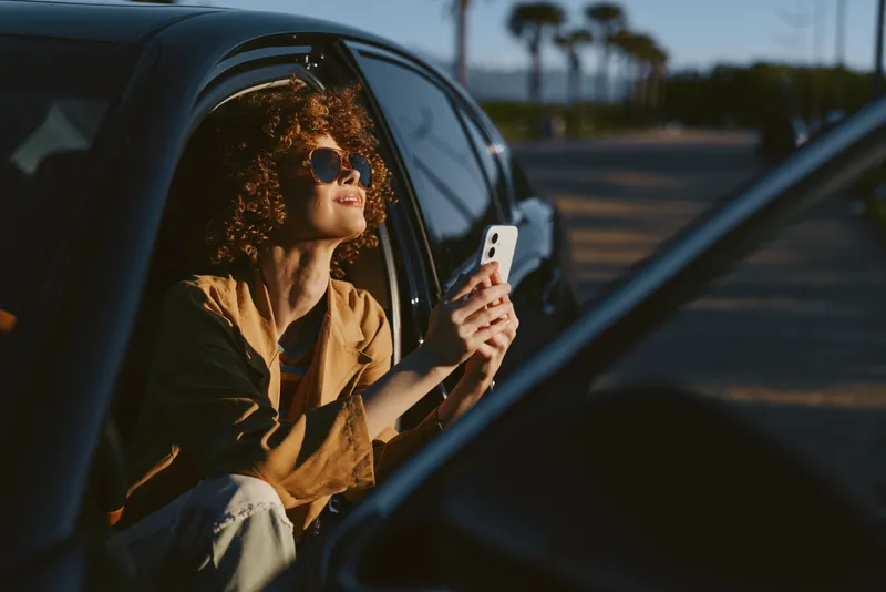 Curly haired woman in a car with sunglasses and phone lifestyle portrait outdoors bright daylight casual fashion warm tones vintage film color confident mood and relaxed vibe