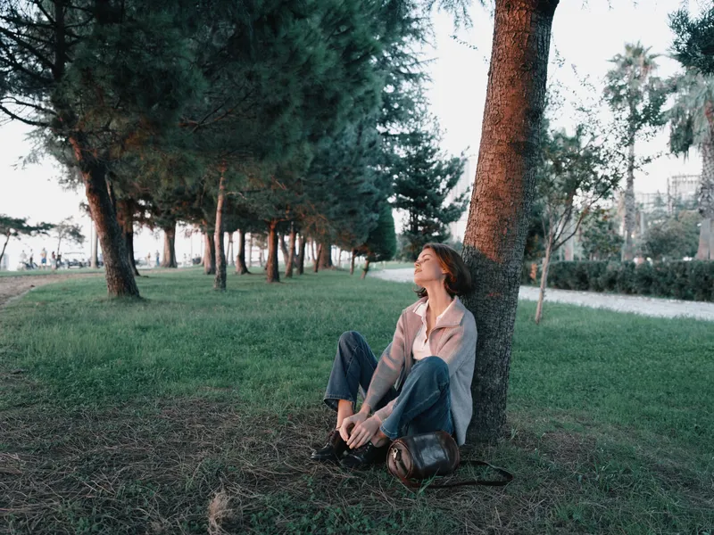 A contemplative young person sits against a tree in a quiet park, gazing upward with a calm, hopeful expression. The scene evokes solitude, reflection, and a peaceful outdoor mood.