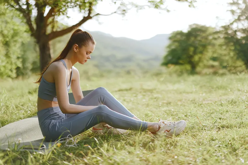 Young athletic woman in blue sportswear sitting on a yoga mat in a lush green field, reflecting serenity and focus with a natural backdrop
