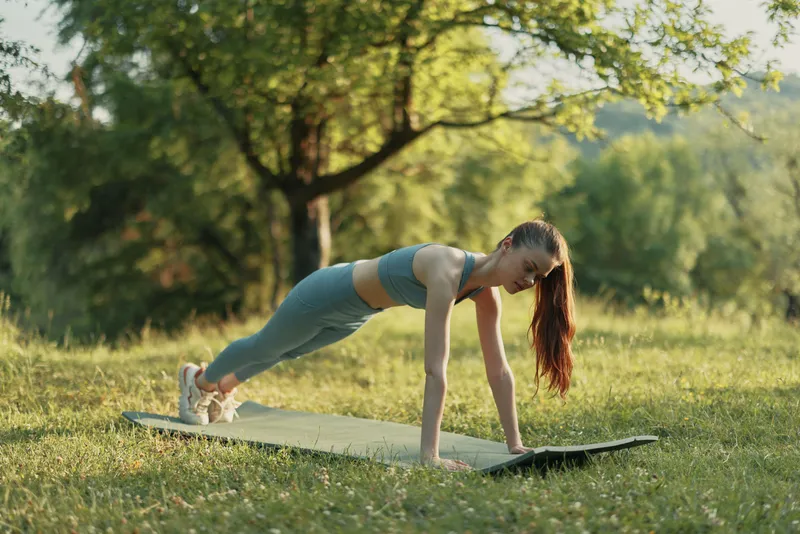 Young athletic woman in sporty outfit performing a plank exercise on a yoga mat outdoors in a lush green park during summer