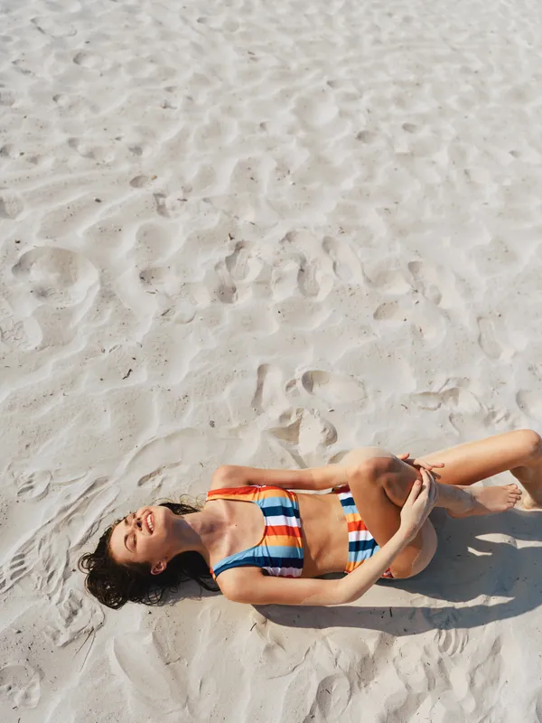A woman lies on sunlit white sand, wearing a colorful striped bikini, relaxed with closed eyes, capturing a bright, carefree summer moment.