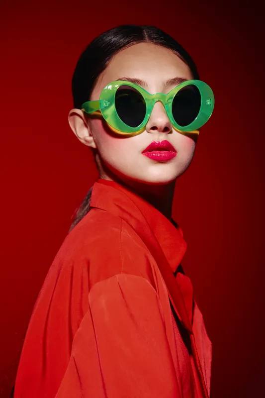 Young woman wearing bright green round sunglasses and vibrant red clothing poses against a red background. Creative fashion and beauty concept with bold colors and unique style.