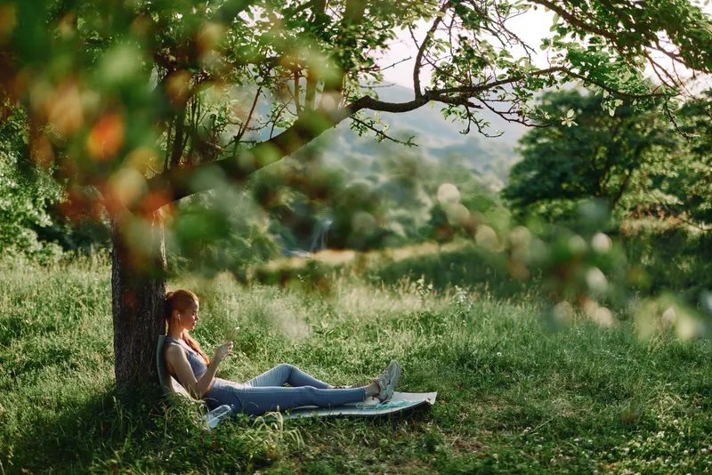 Young woman relaxing outdoors under a tree, enjoying nature and reading a book in a serene green landscape, conveying tranquility and inspiration