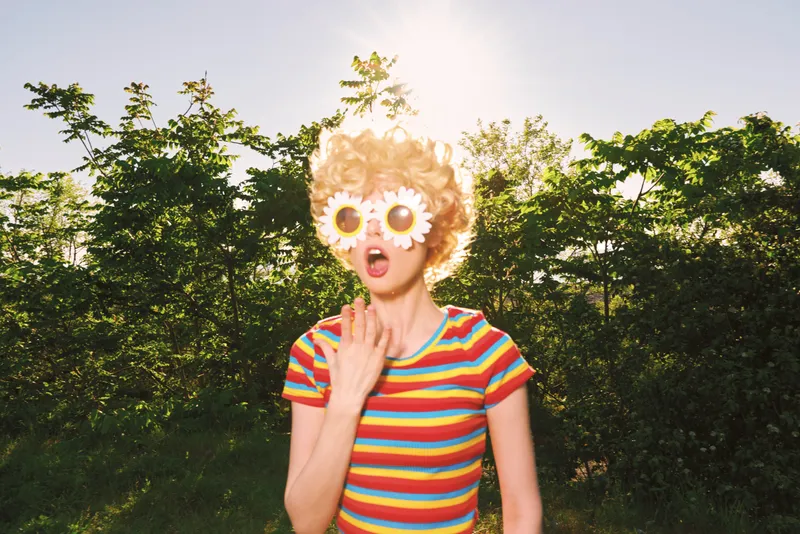 Joyful woman outdoors wearing a striped shirt and playful sunflower sunglasses, captured in retro film color, conveying carefree summer mood amid lush trees and clear skies above