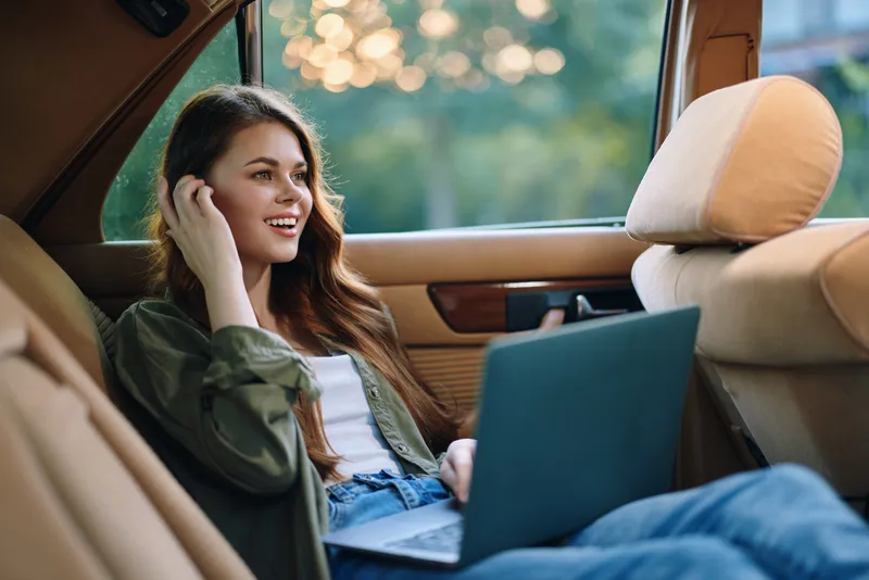 Young woman enjoying the outdoors while working on a laptop inside a vintage car, presenting a relaxed and joyful lifestyle amidst nature.