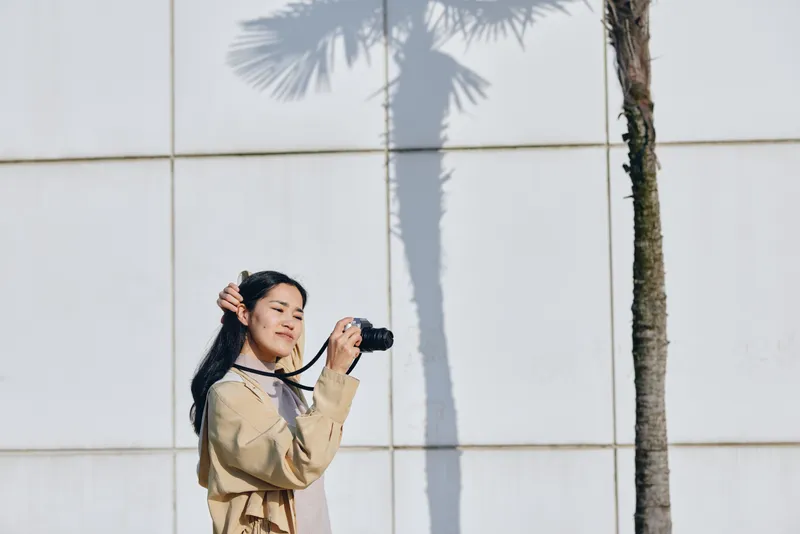 Woman photographer with camera in hand stands outdoors beside a white wall, palm tree shadow stretching across the ground, wearing a beige jacket, capturing a candid street moment under bright light
