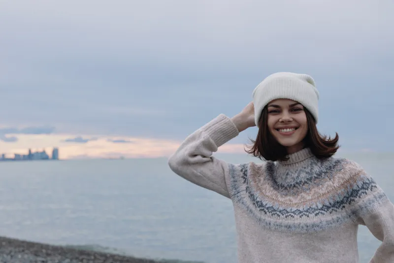 Woman in a cozy knit sweater and beanie smiles at the sea, outdoors by the shore during cool weather, capturing a relaxed and warm coastal moment.