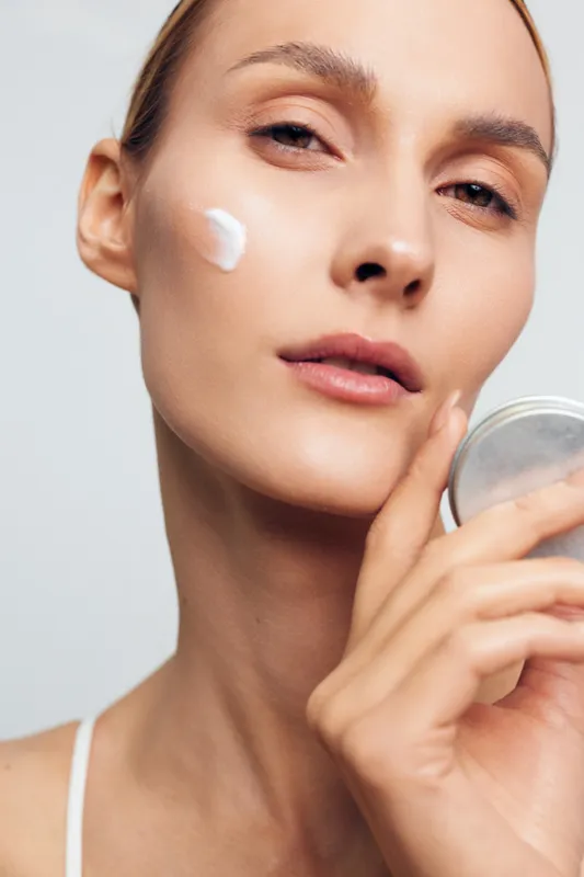 A young woman with clear skin applies white cream on her cheek while holding a container in a bright studio. Concept for skincare, beauty routine, and natural complexion care.