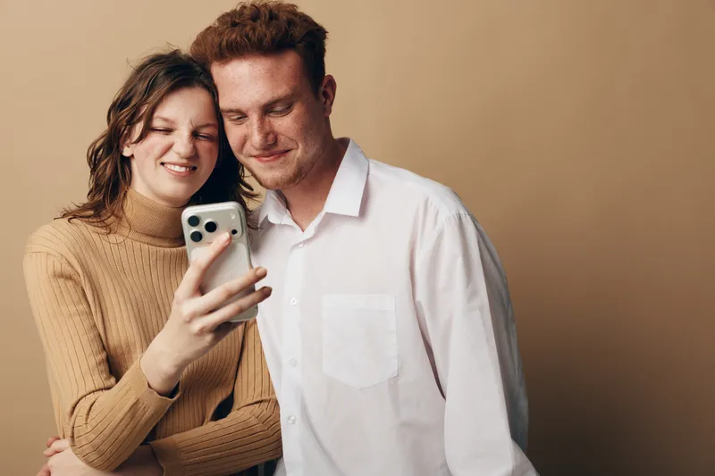Smiling young couple looking at smartphone together, standing close with warm beige background. Concept of technology, relationship, communication, and modern lifestyle.