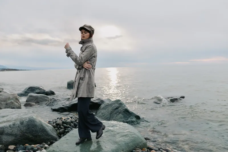 Young woman in coat and hat stands on rocky beach near calm sea during cloudy sunset. Fashion outdoor portrait with serene water background and soft natural light.