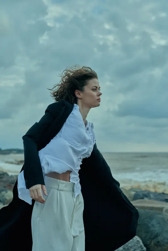 Windy beach scene with a flowing coat, white shirt and light pants, dynamic pose against stormy sky and sea for fashion and coastal mood.