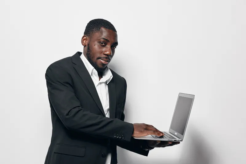 African American man with dark skin wearing black suit and white shirt working on laptop computer standing against white background. Business professional using technology for work.