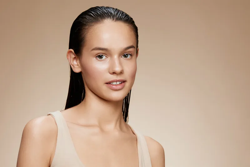 Natural beauty portrait of a young woman with wet hair, showcasing healthy skin and a neutral expression against a soft beige background