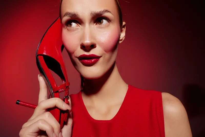 Close-up of a woman in red dress holding a red high heel shoe near her face against a red background. Concept of fashion, style, beauty, and bold color contrast in studio lighting.