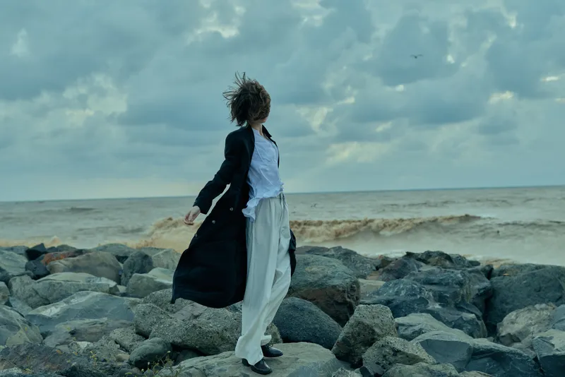 Windy shoreline scene with flowing coat and loose trousers on a rocky beach, ocean horizon and dramatic sky conveying motion and solitude