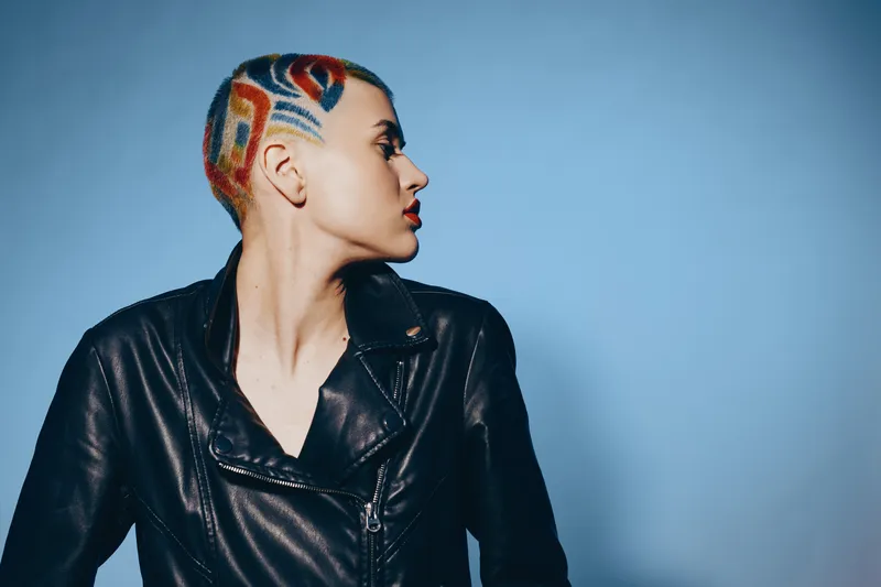 Young woman with colorful shaved hairstyle wearing black leather jacket looking sideways against blue background, showcasing bold and artistic fashion style.