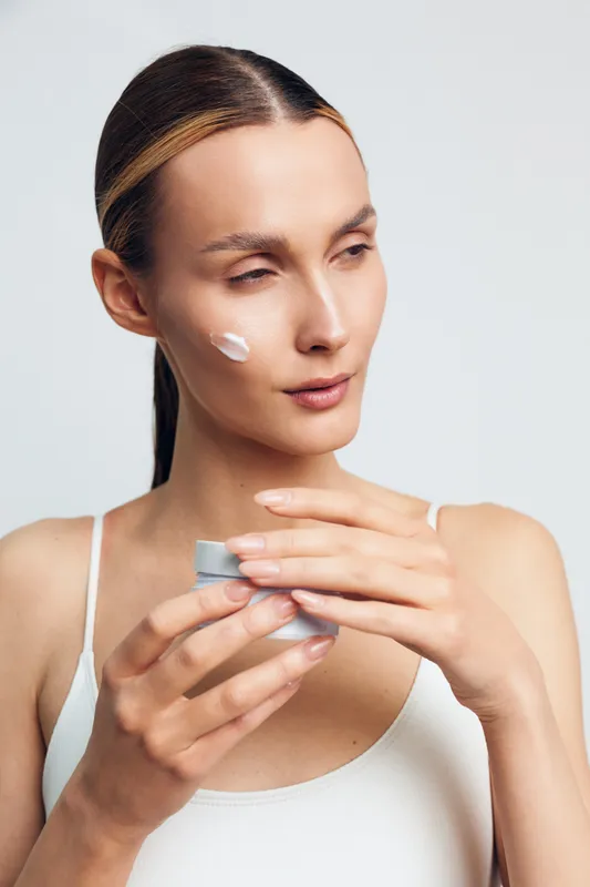 Young woman with smooth skin applies face cream from small jar while looking away isolated on white background with natural makeup and focused expression.